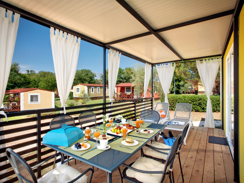 Overdekt terras bij Maravea Family Holiday Home met eettafel, witte gordijnen en uitzicht op chalets.