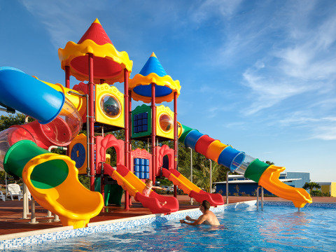 Scivoli colorati vicino a una piscina, con un bambino che gioca con un adulto sotto un cielo sereno.