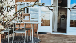 Holzterrasse am Smûk Fjildhuske mit Tisch und Stühlen, blühende Zweige und moderne Lodge im Hintergrund.