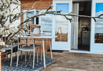 Wooden patio at Smûk Fjildhuske lodge, with outdoor dining set, blooming branches and cozy cabin view.