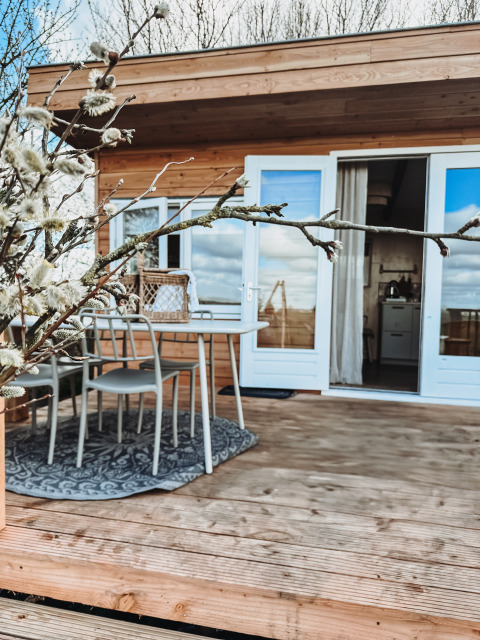 Terraza de madera en Smûk Fjildhuske, con mesa, sillas, ramas florecidas y lodge acogedor de fondo.