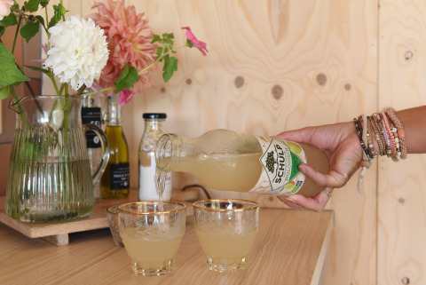 Una persona sirve jugo en vasos sobre una mesa de madera en una acogedora tiny house con flores y bebidas.