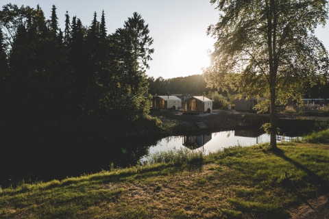 Foto van de Wrap house lodge met sauna en badkuip bij een vijver, omringd door bomen en zonlicht.