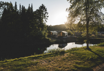 Foto van de Wrap house lodge met sauna en badkuip bij een vijver, omringd door bomen en zonlicht.