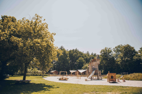 Spielplatz mit Holzspielgeräten und Rutsche am Safarizelt im Veluwepark de Bosgraaf, Niederlande, unter Bäumen.