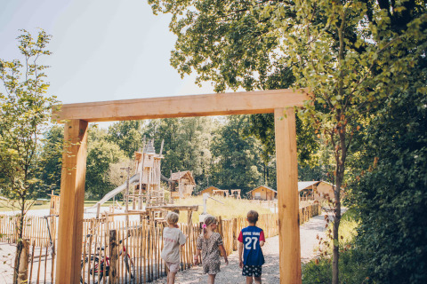 Drei Kinder betreten einen Spielplatz bei einer Glamping-Unterkunft, umgeben von Bäumen und Holzhütten.