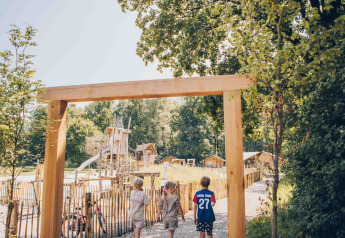 Tre bambini entrano in un'area giochi in legno presso una sistemazione glamping circondata da alberi e casette.