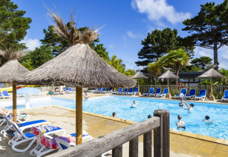 Außenpool im Flower Camping Port de la Chaîne in der Bretagne, Frankreich, mit Sonnenliegen und Strohdächern.