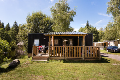 Holzhütte mit Veranda am Flower Camping La Sténiole in Grand Est, Frankreich, von Bäumen umgeben.