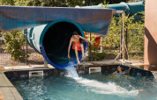 Un garçon sort d’un toboggan aquatique dans la piscine, observé par une fille, Flower Camping La Sténiole, Grand Est, France.