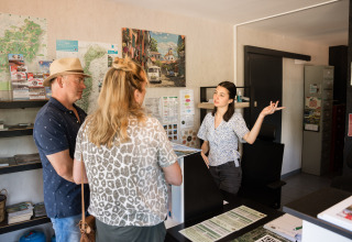 Twee bezoekers krijgen uitleg van een receptioniste bij Flower Camping La Sténiole in Grand Est, Frankrijk.