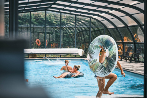 Piscine couverte à Flower Camping La Sténiole avec des vacanciers jouant et se relaxant sur des bouées gonflables.