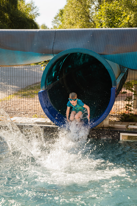 Un bambino esce da uno scivolo blu e si tuffa in piscina al Flower Camping La Sténiole, Grand Est, Francia.