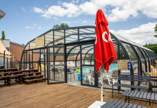 Piscina coperta da vetro, ombrellone rosso e terrazza in legno al Flower Camping La Sténiole, Grand Est, Francia.