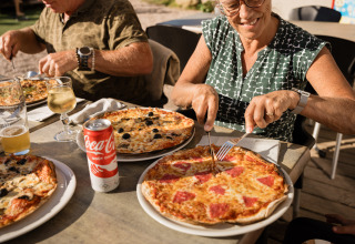 Two people enjoy pizza and drinks outdoors at Flower Camping La Sténiole, Grand Est, France holiday park.