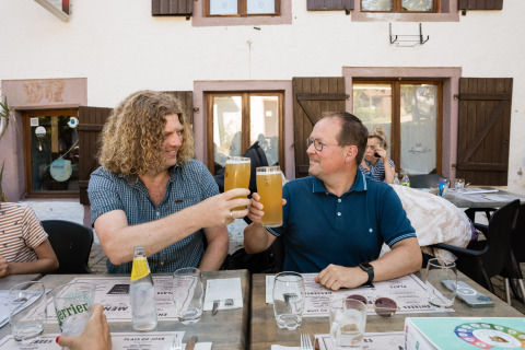 Deux hommes trinquent avec de grandes bières en terrasse au Flower Camping La Sténiole, Grand Est, France.