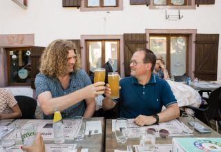 Deux hommes trinquent avec de grandes bières en terrasse au Flower Camping La Sténiole, Grand Est, France.
