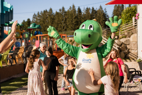 Kinder genießen Aktivitäten mit einem grünen Dinosauriermaskottchen im Flower Camping La Sténiole, Frankreich.