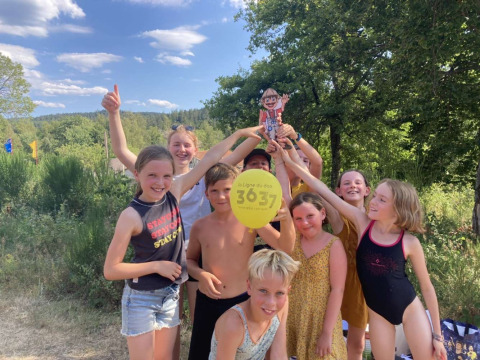 Eine fröhliche Kindergruppe hält einen Ballon und eine Pappfigur im Grünen bei sonnigem Wetter hoch.