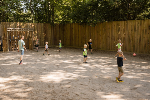 Children play soccer on a sandy field with a wooden fence at Flower Camping La Sténiole in Grand Est, France.