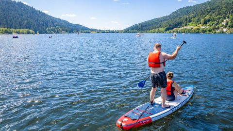 To personer står på et paddleboard på en sø nær Granges-Aumontzey, omgivet af grønne bakker.