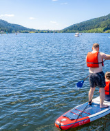 To personer står på et paddleboard på en sø nær Granges-Aumontzey, omgivet af grønne bakker.