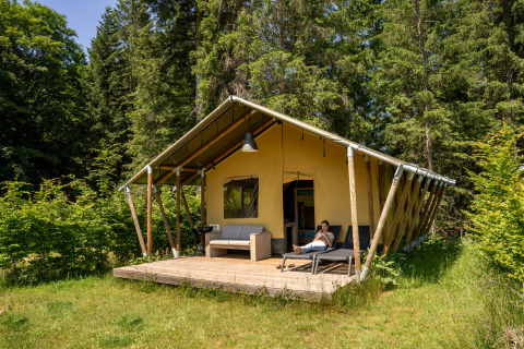 Safari-telt ved Flower Camping La Sténiole i Frankrig, omgivet af skov og en afslappet gæst på terrassen.