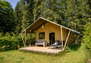 Safari-telt ved Flower Camping La Sténiole i Frankrig, omgivet af skov og en afslappet gæst på terrassen.