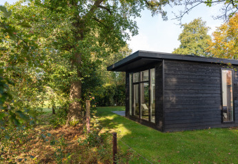 Sort træ-lodge kaldet Wellness Boutique baby, omgivet af grønne træer og græsplæne i solskin.