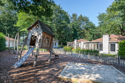 Ein Spielplatz mit Holzhütte und Rutsche steht vor modernen Glamping-Unterkünften im Grünen.