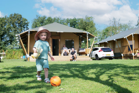 Jeune enfant joue au ballon devant une tente safari de luxe, parents détendus sur la terrasse.
