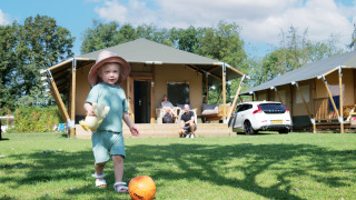 Bambino gioca con la palla davanti a una tenda safari di lusso, genitori rilassati sulla terrazza.