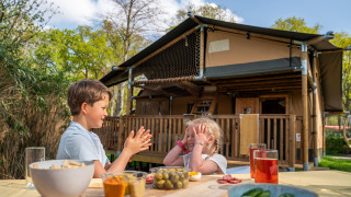 Two children play at a table with snacks in front of a Rangerlodge safari tent in a sunny setting.