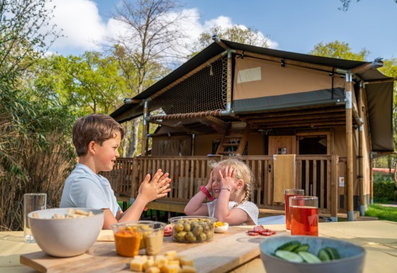 Two children play at a table with snacks in front of a Rangerlodge safari tent in a sunny setting.