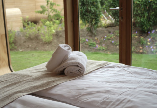 Udsigt fra Wrap House-lodge på Resort de Parel i Holland med håndklæder på seng og grøn have udenfor.