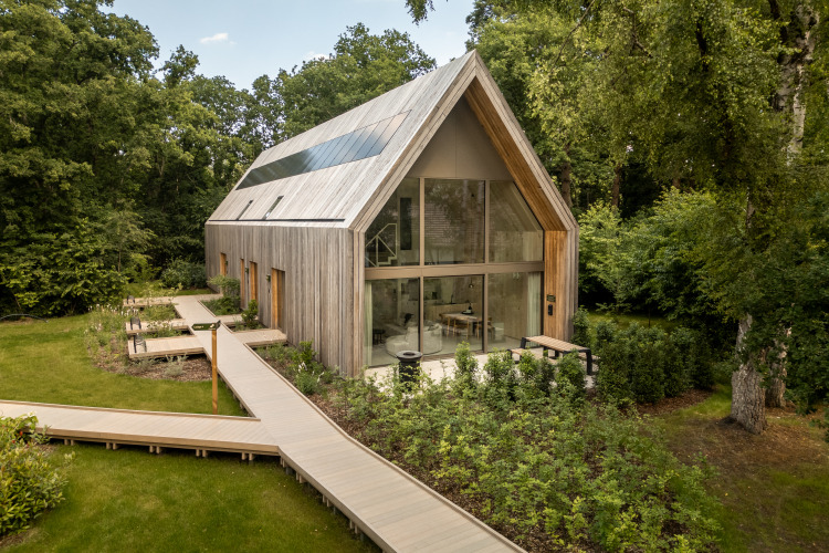 Lodge moderno de madera con grandes ventanales, rodeado de bosque y pasarela de madera en el jardín.
