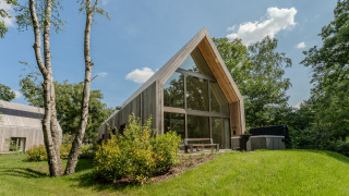 Moderne Lodge mit großen Glasfenstern und Jacuzzi, umgeben von grüner Natur und blauem Himmel.