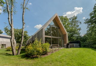 Moderne lodge met grote glaspartijen en jacuzzi, omgeven door natuur en bomen op een zonnige dag.