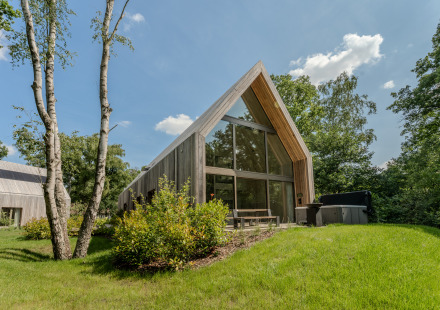 Moderne lodge met grote ramen en jacuzzi, omringd door groen en bomen onder een zonnige hemel.