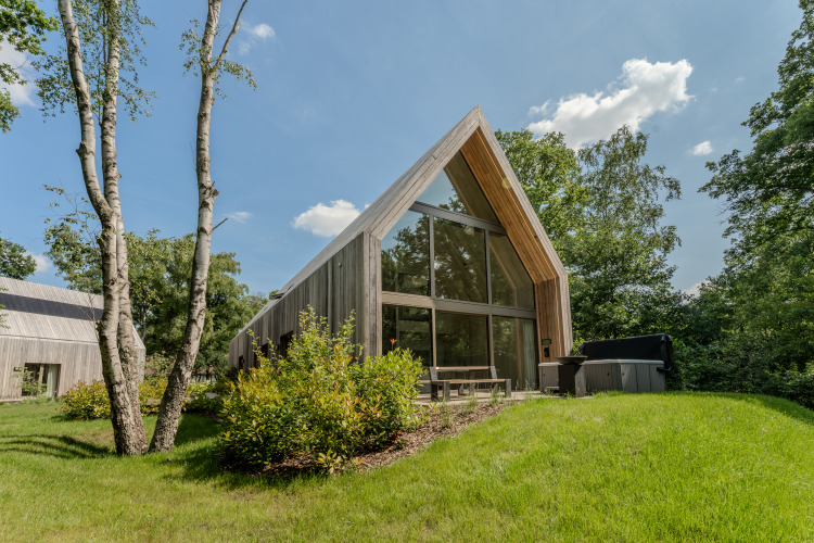 Lodge moderno con grandes ventanales y jacuzzi, rodeado de naturaleza y césped bajo un cielo soleado.