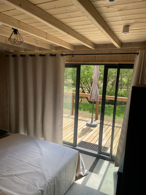 Chambre avec plafond en bois, grandes rideaux et vue sur terrasse au Cocoon de l’Ourthe, Hameau de l’Ourthe, Belgique.