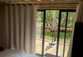 Chambre avec plafond en bois, grandes rideaux et vue sur terrasse au Cocoon de l’Ourthe, Hameau de l’Ourthe, Belgique.
