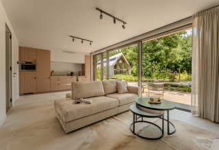 Helles Wohnzimmer einer Lodge mit großen Fenstern, offener Küche und Blick auf einen grünen Garten.