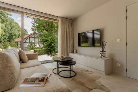 Moderne lodge-stue med stor sofa, tv på væggen, glasdør og udsigt til haven og bindingsværkshus.
