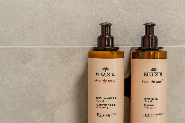 Dos botellas de Nuxe Réve de Miel con champú y acondicionador montadas en una pared de azulejos en un lodge.