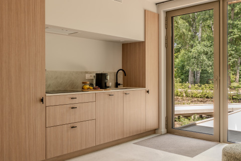 Pequeña cocina moderna de madera clara junto a un gran ventanal con vista al bosque en una cabaña.