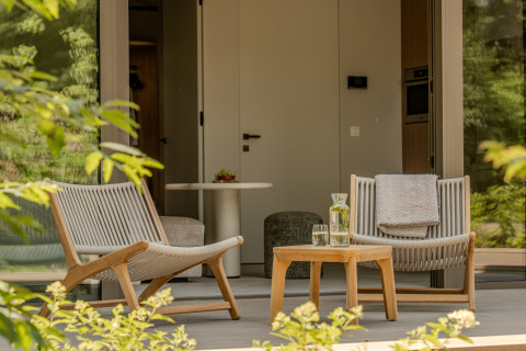 Dos sillones modernos y una mesa con agua y vasos en un porche de lodge acogedor rodeado de vegetación.