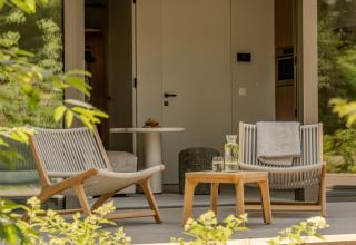 Dos sillones modernos y una mesa con agua y vasos en un porche de lodge acogedor rodeado de vegetación.