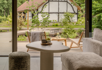 Hyggeligt lodge-interiør med udsigt til en terrasse og traditionelt hus omgivet af grønne træer og planter.