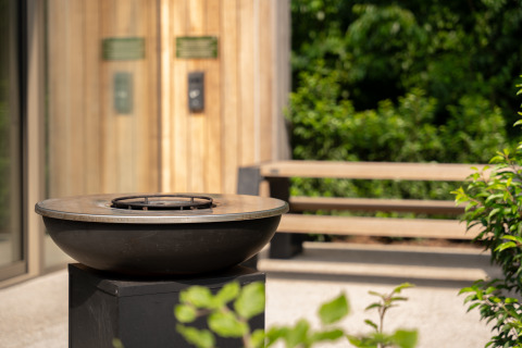 Moderne buitenvuurkorf bij Lodge, omringd door groene planten, bank en houten wand.
